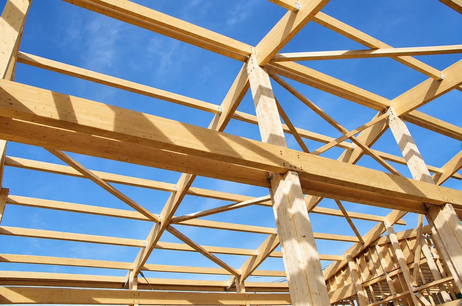 Structure de charpente en bois clair composée de poutres et de poteaux assemblés, photographiée en contre-plongée sur un ciel bleu.